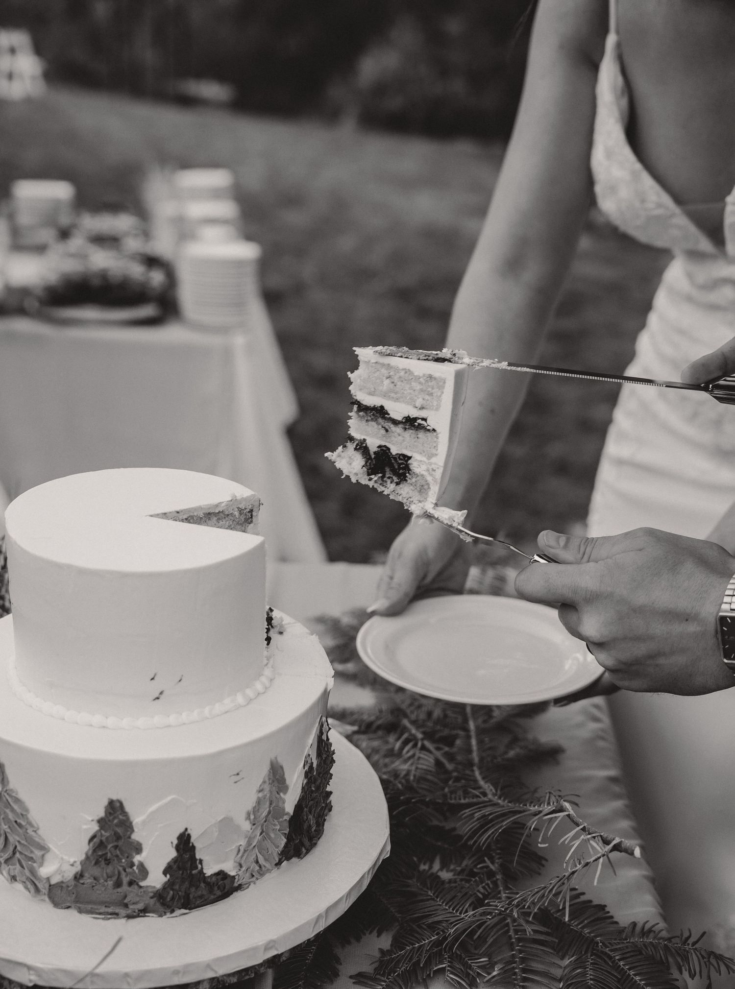 A white two-tier wedding cake being cut, decorated with a hand-painted forest scene.
