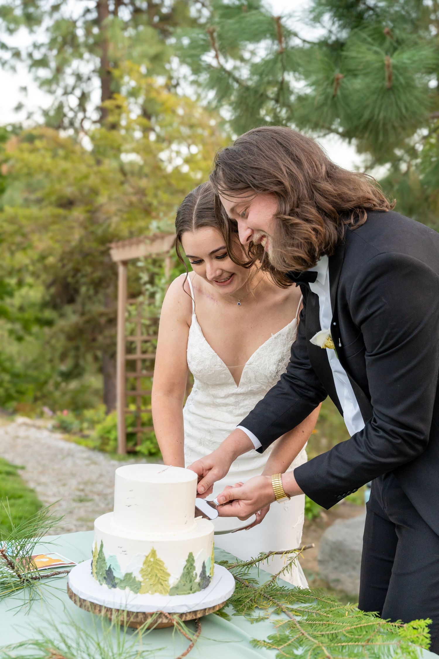 A couple cuts their white wedding cake decorated with pine trees in an outdoor garden setting.