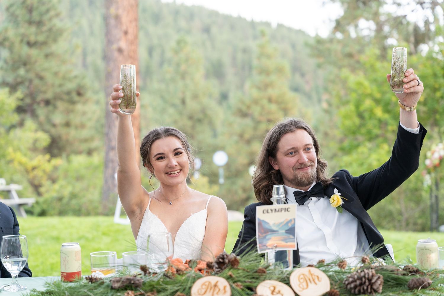 A wedding couple raises their glasses in celebration at an outdoor reception table decorated with rustic decor.