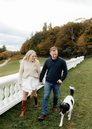 Couple walking with black and white dog along white balustrade in autumn setting.