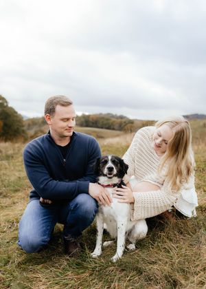 Couple sits in grassy field with their small black and white dog during outdoor photoshoot.