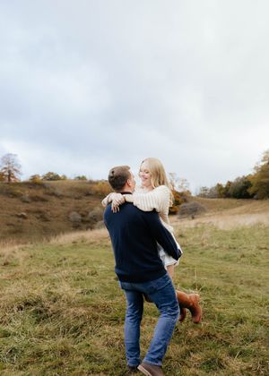 Couple sharing playful moment on hillside with cloudy sky background.