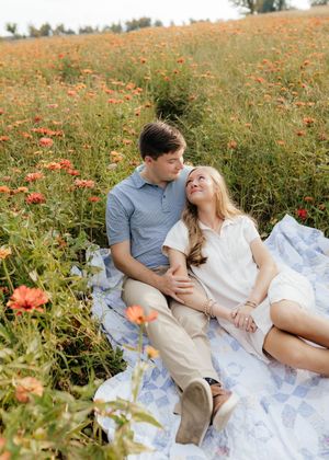 Couple lounges on white blanket amid blooming orange wildflowers during golden hour.