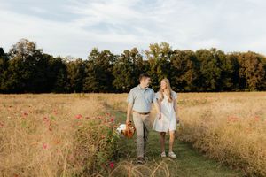 Couple walking hand in hand through a golden meadow with red wildflowers and autumn trees.