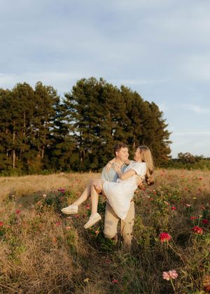 Romantic couple shares playful moment in wildflower meadow with pine trees at sunset.