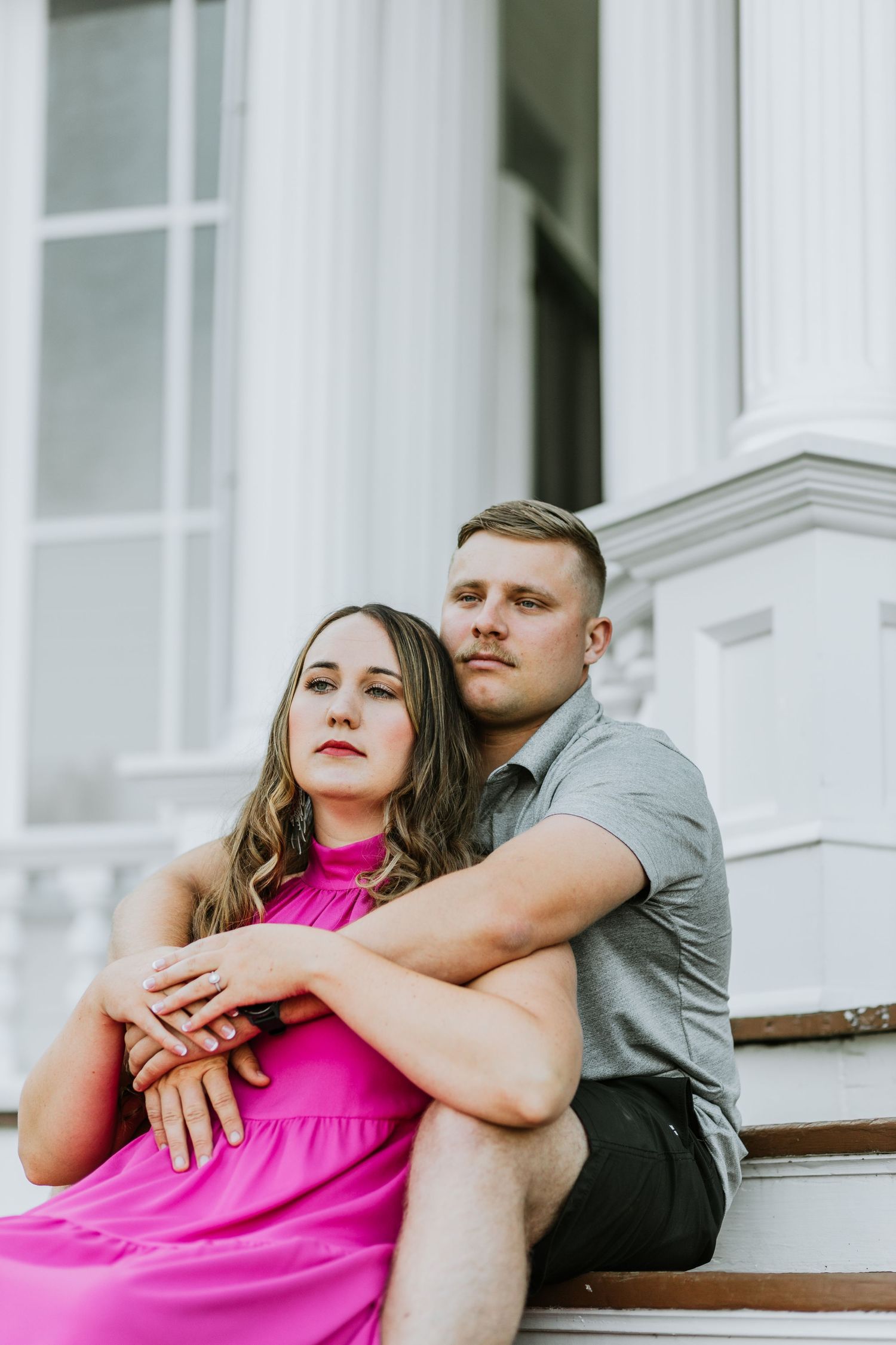 A couple poses intimately on white steps wearing pink and gray casual attire.
