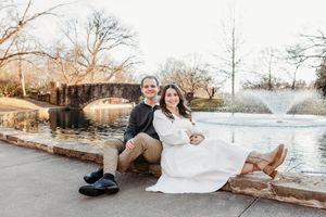 A romantic winter photoshoot by a lake with bare trees and a stone bridge in the background.