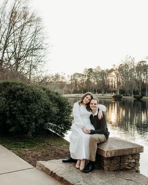 A couple sits together on a stone bench beside a tranquil lake at sunset.
