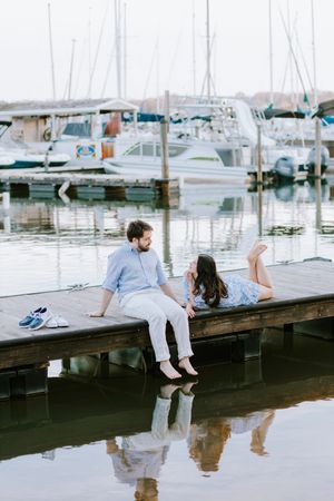 A couple relaxes on a wooden dock with boats and their reflection visible in the calm water.