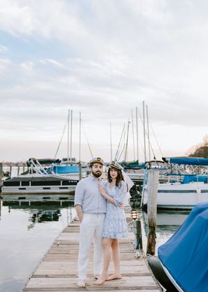 A couple in matching light outfits stands on a marina dock surrounded by sailboats.
