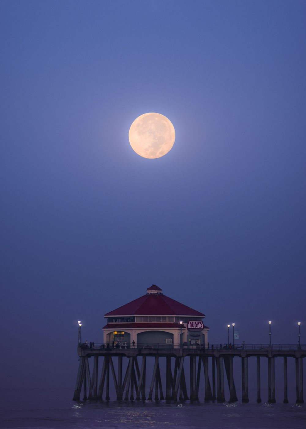 Full moon glows above Huntington Beach Pier at night, reflecting off the ocean and silhouetting the structure, captured by Chris Cornish Pho