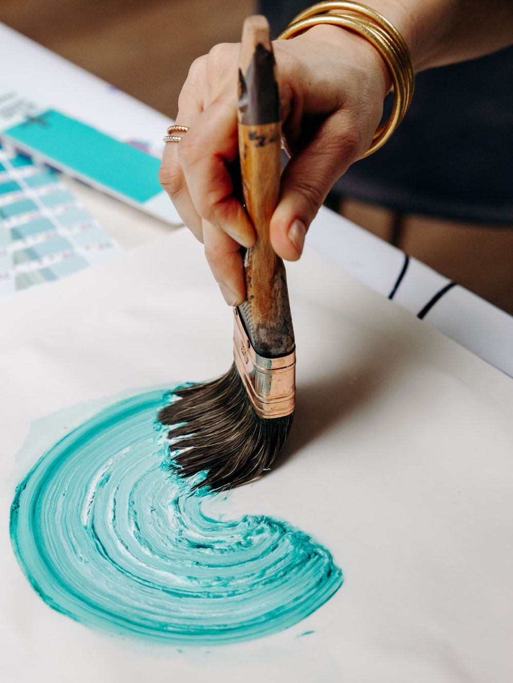 A hand holding a worn paintbrush creates a teal swirl on white paper, with gold bangles and rings visible.