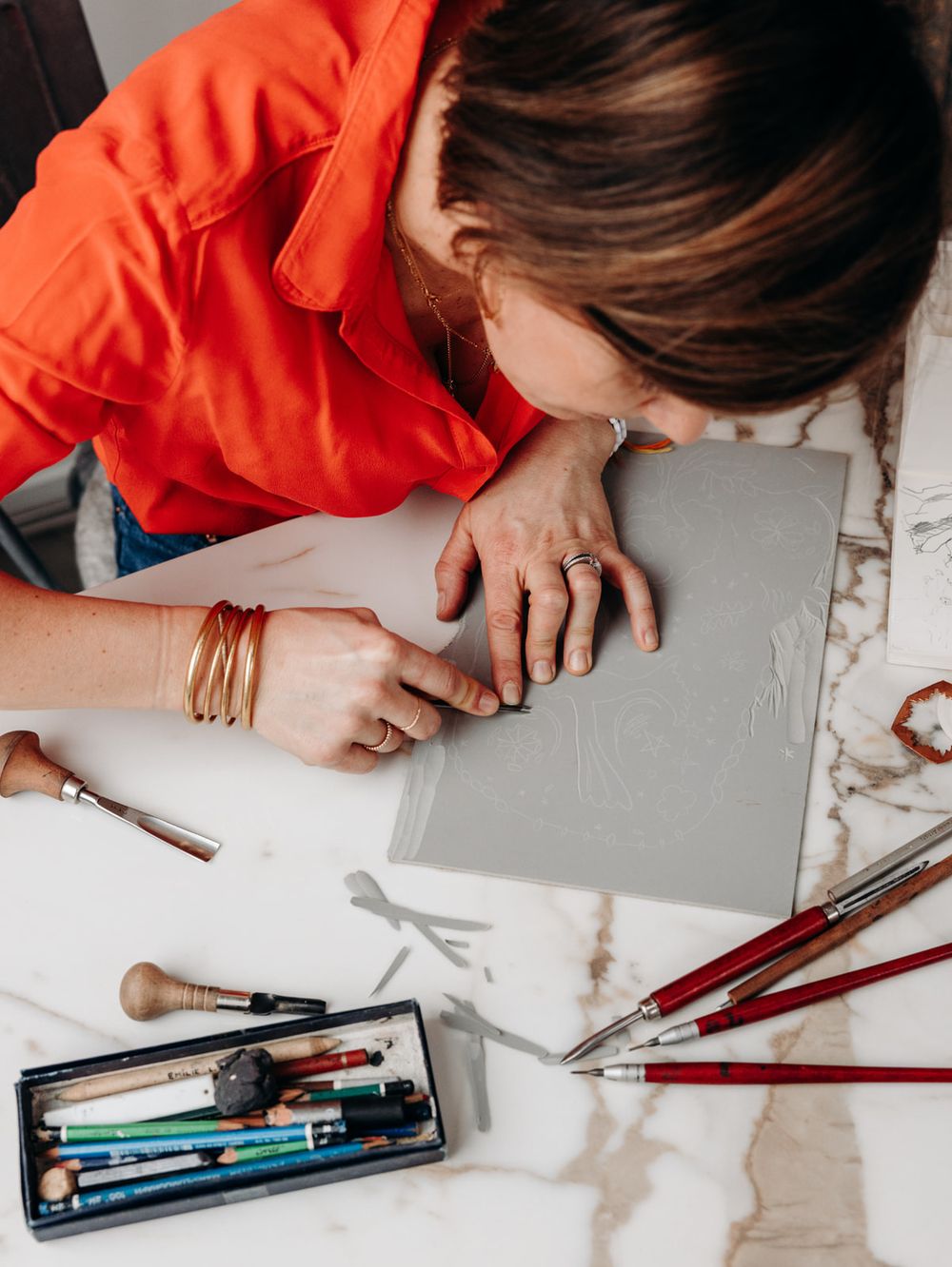 Artist in orange shirt works on gray canvas with brushes and art tools on marble table.