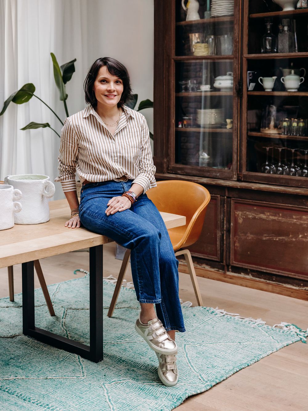 Woman in striped shirt and jeans sits on wooden table beside amber chair and vintage cabinet.