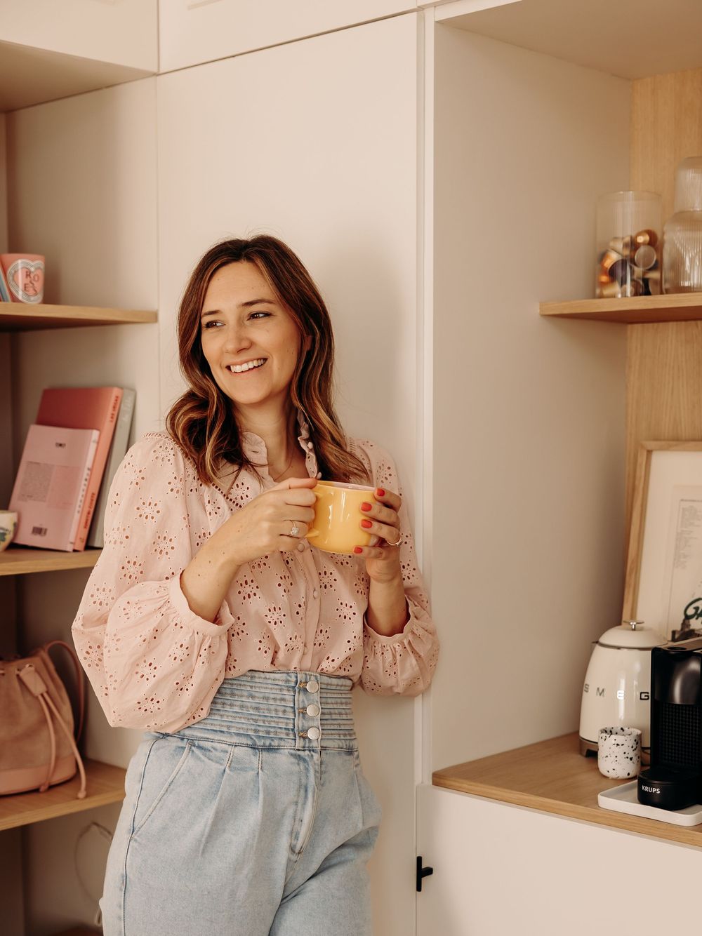 Smiling woman in pink blouse holding yellow mug in a stylish home office with shelves.