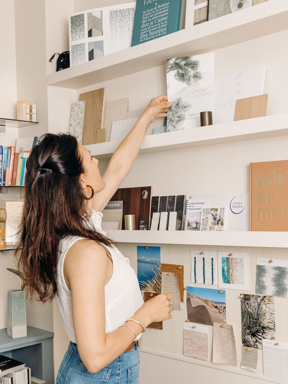 Woman reaching up to arrange books and decor on white wall shelves in a bright, modern interior design studio.