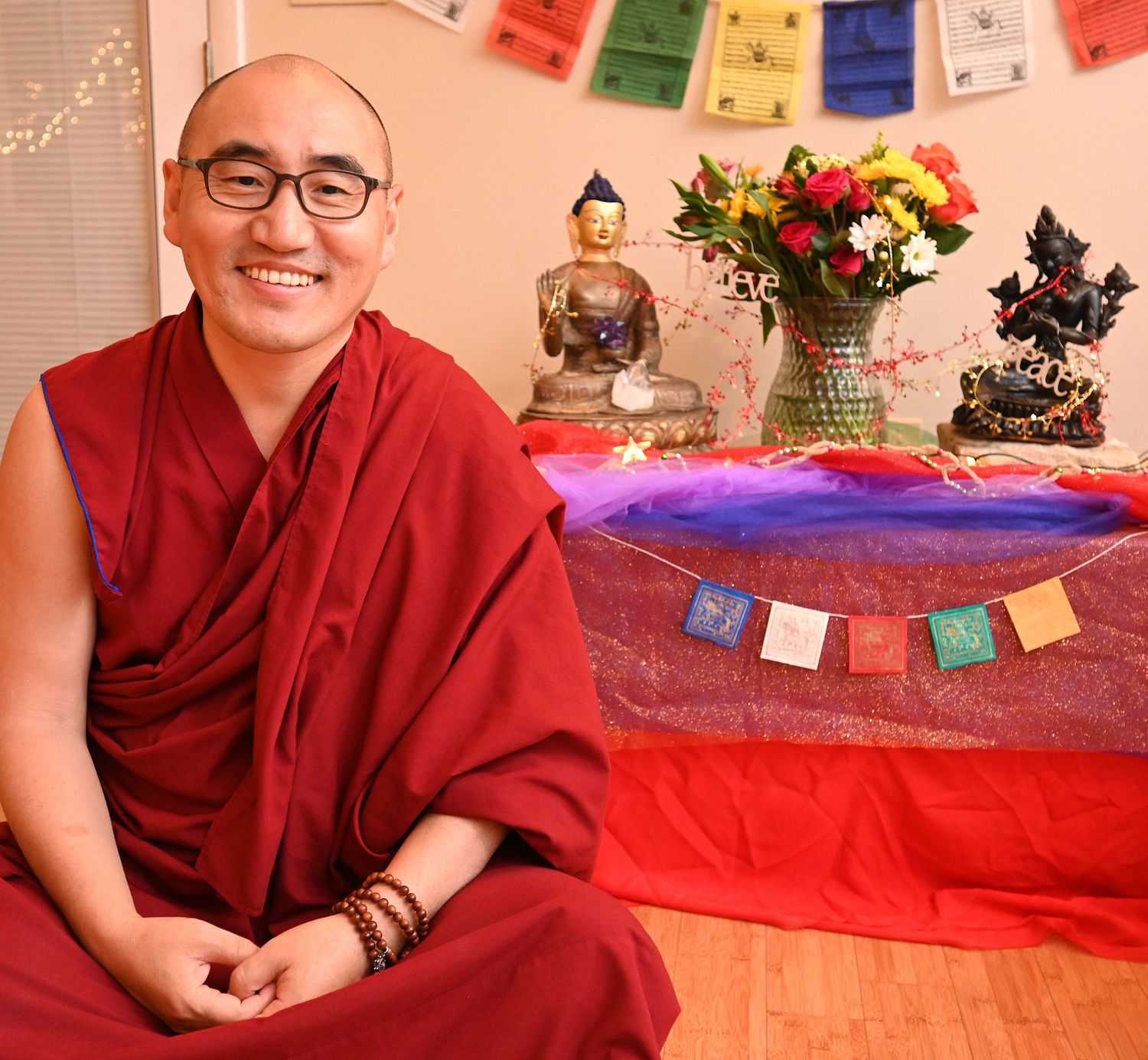 Buddhist monk in traditional red robes sitting by altar with flowers and decorative flags | photo by Tom McCall Photography | Maryland Photo