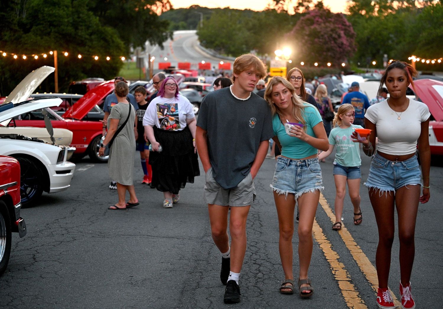 People enjoying a summer evening at a classic car show with string lights and sunset in the background.