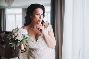 An elegant bride in a lace wedding dress holds a pink and white bouquet while standing by a window.