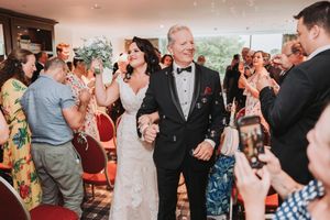 Wedding guests cheer and take photos as a couple exits their ceremony in a beautiful venue with floor to ceiling windows.