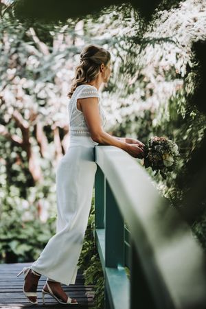 Person in a white dress stands on a wooden deck surrounded by lush greenery and soft sunlight.