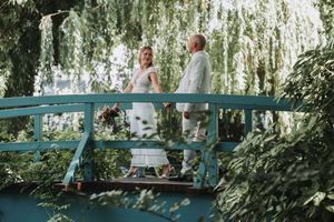 A couple walks hand in hand through a lush garden path with hanging willow trees and dappled sunlight.