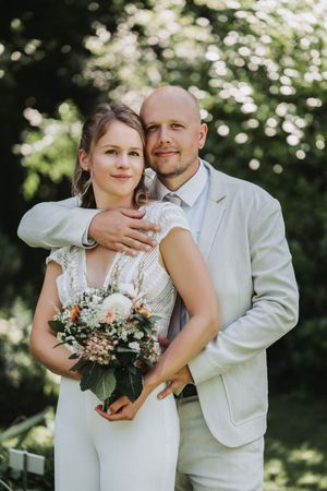 A romantic wedding couple embraces outdoors in natural lighting with a floral bouquet and light-colored attire.
