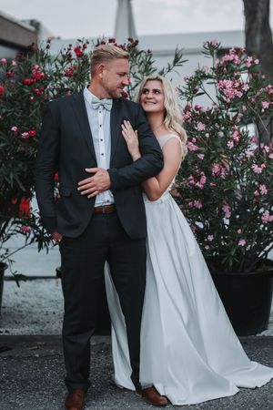 A romantic wedding couple embraces near blooming pink and red oleander bushes in an elegant outdoor setting.