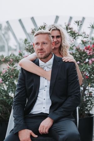 A couple poses together outdoors for a romantic wedding portrait with flowering plants in the background.