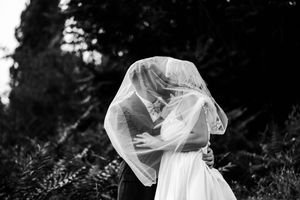 A bride and groom embrace in a romantic outdoor photoshoot surrounded by lush greenery and trees.