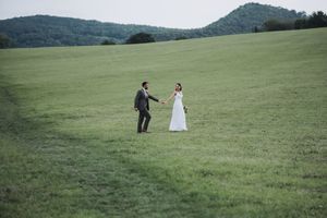 Wedding couple walks across a scenic green meadow with rolling hills in the background.