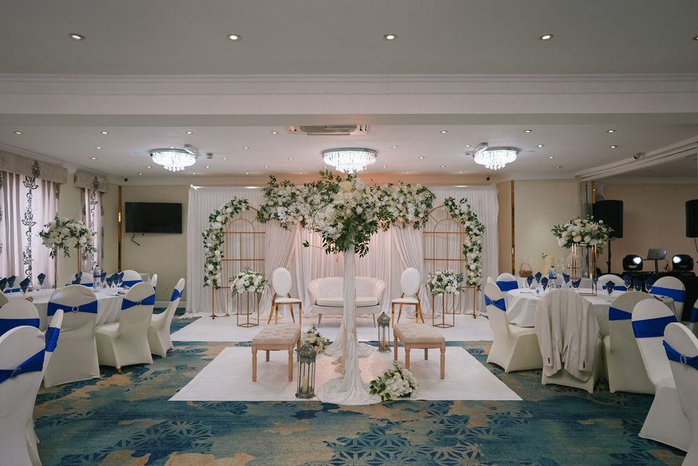 Wedding reception venue decorated in white and blue with round tables, floral centerpieces, and elegant ceiling lighting.