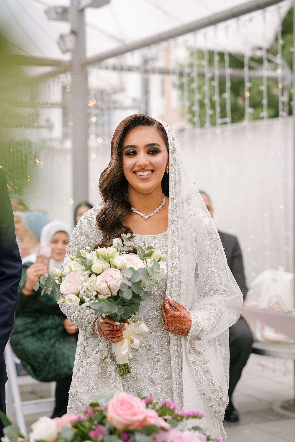 Bride in flowy lace veil holds a romantic bouquet of pink and white flowers during an outdoor wedding ceremony.
