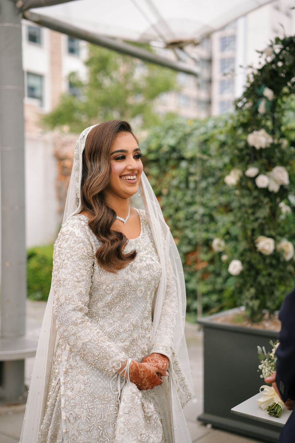 A bride in a detailed white lace dress and veil poses outdoors against a backdrop of greenery and roses.