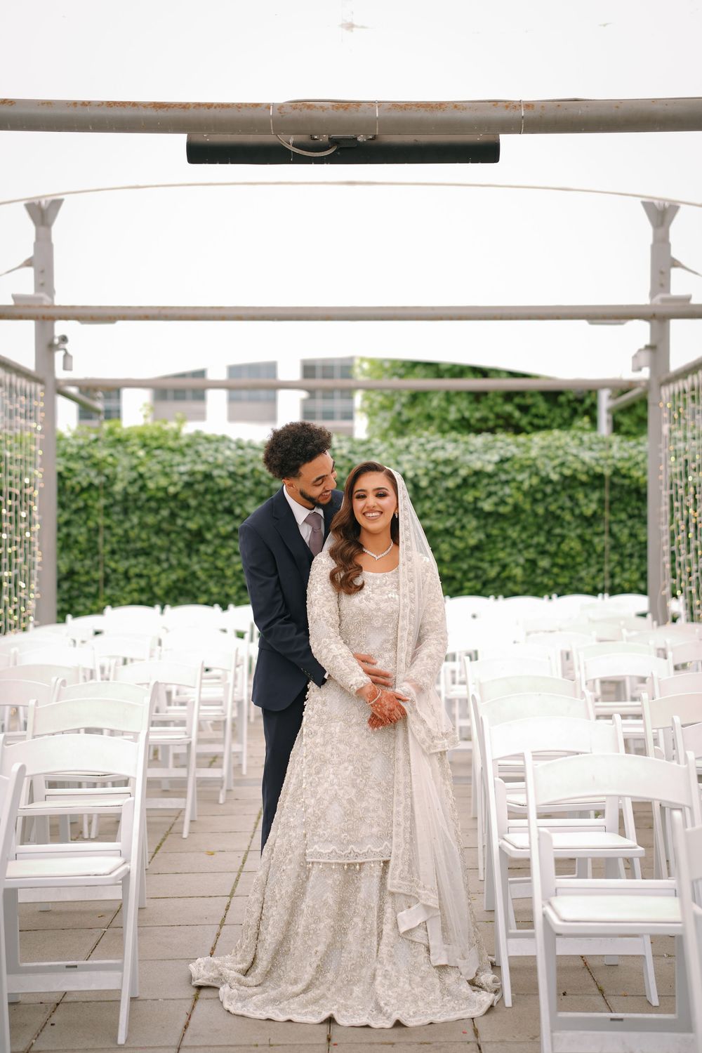 A couple stands together at their outdoor wedding venue with white chairs arranged for the ceremony.