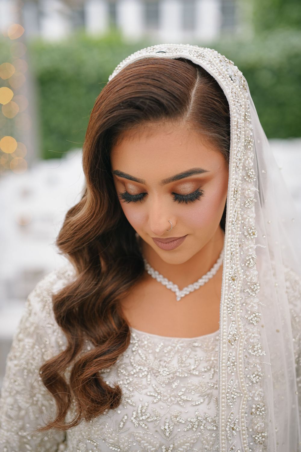 Bride wearing delicate lace veil and crystal necklace with flowing brown curls and soft glam makeup looking down.