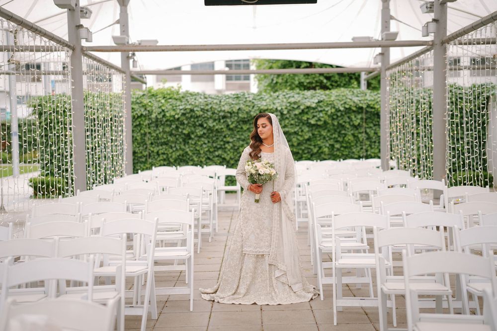 A wedding ceremony setup with white chairs and hanging string lights in a modern outdoor space with greenery walls.