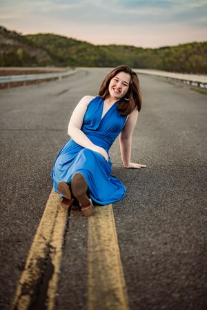 Person in a blue dress poses playfully on a road's yellow lines at dusk while surrounded by hills and nature.