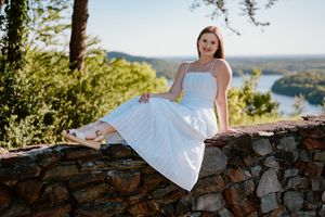 Young woman in white sundress sitting on stone wall overlook with scenic river and forest views behind her.