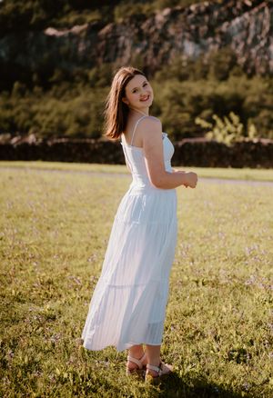 Young woman in a white midi dress smiling over her shoulder in a sunlit green meadow with rocky hills behind her.