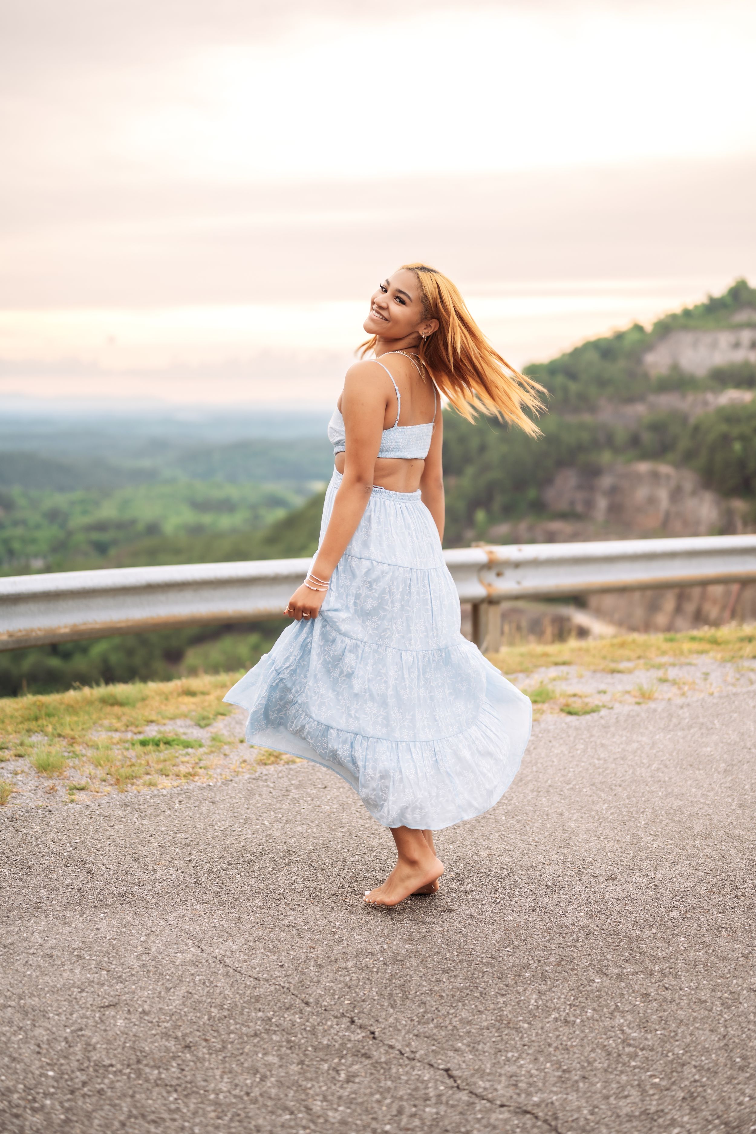Young woman in light blue crop top and maxi skirt twirling on mountain road at sunset.