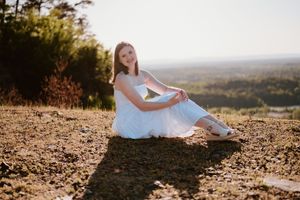 Young woman in white dress sitting on a hillside with scenic countryside views in golden hour sunlight.