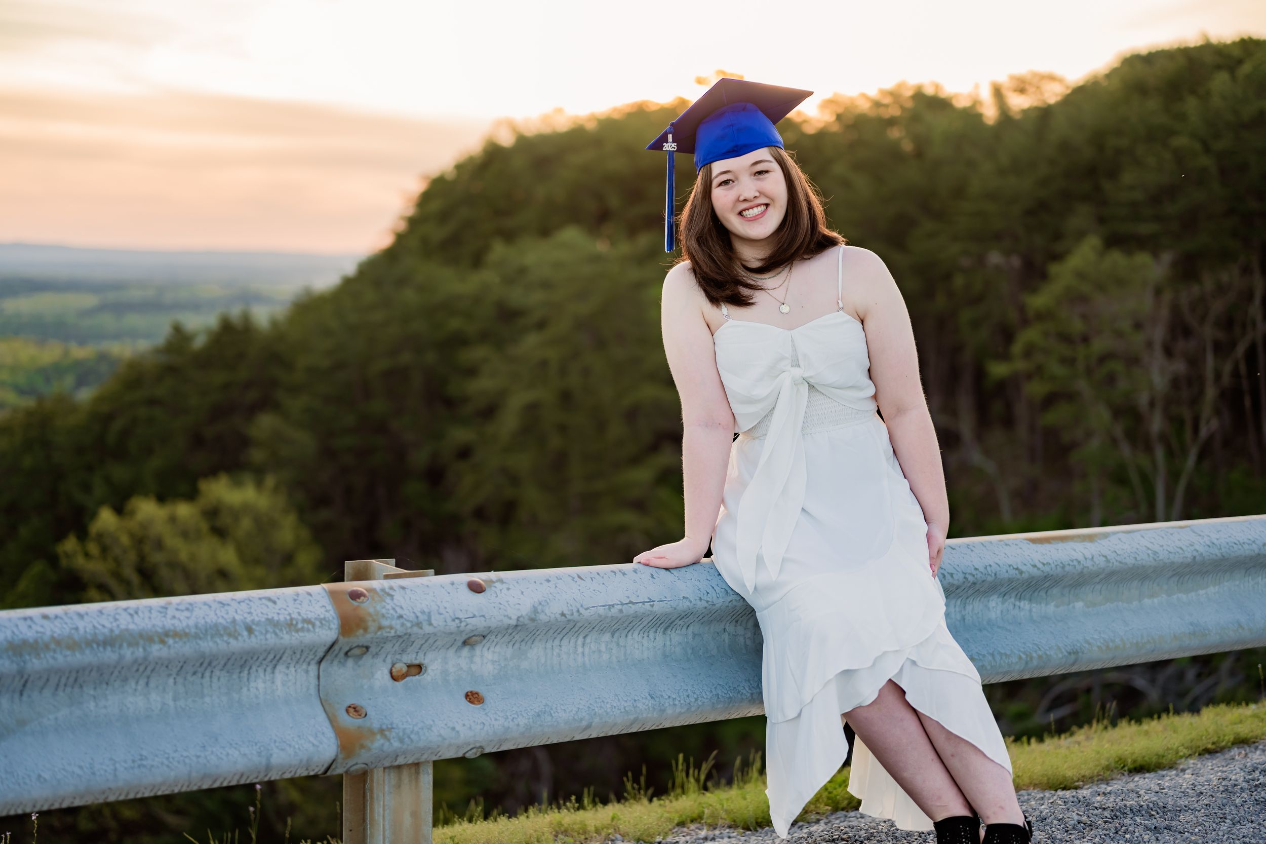 Graduate in blue cap and gown stands against scenic mountain landscape at sunset.