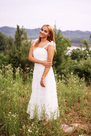 Young woman in white lace maxi dress standing in green field with mountains and trees in background.