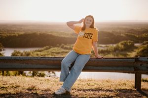 Young woman in yellow State shirt and jeans leaning on a fence railing at golden hour with scenic landscape behind her.