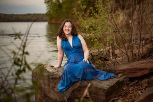 A figure in a flowing royal blue dress poses elegantly on wooden steps near a serene lake at dusk.