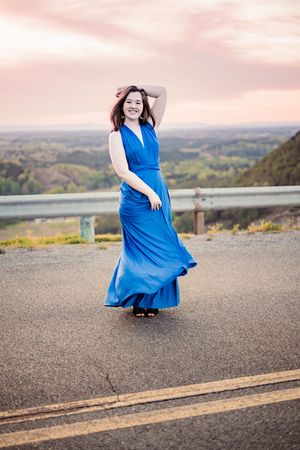 A person in a flowing blue maxi dress poses expressively on a scenic mountain overlook at sunset.