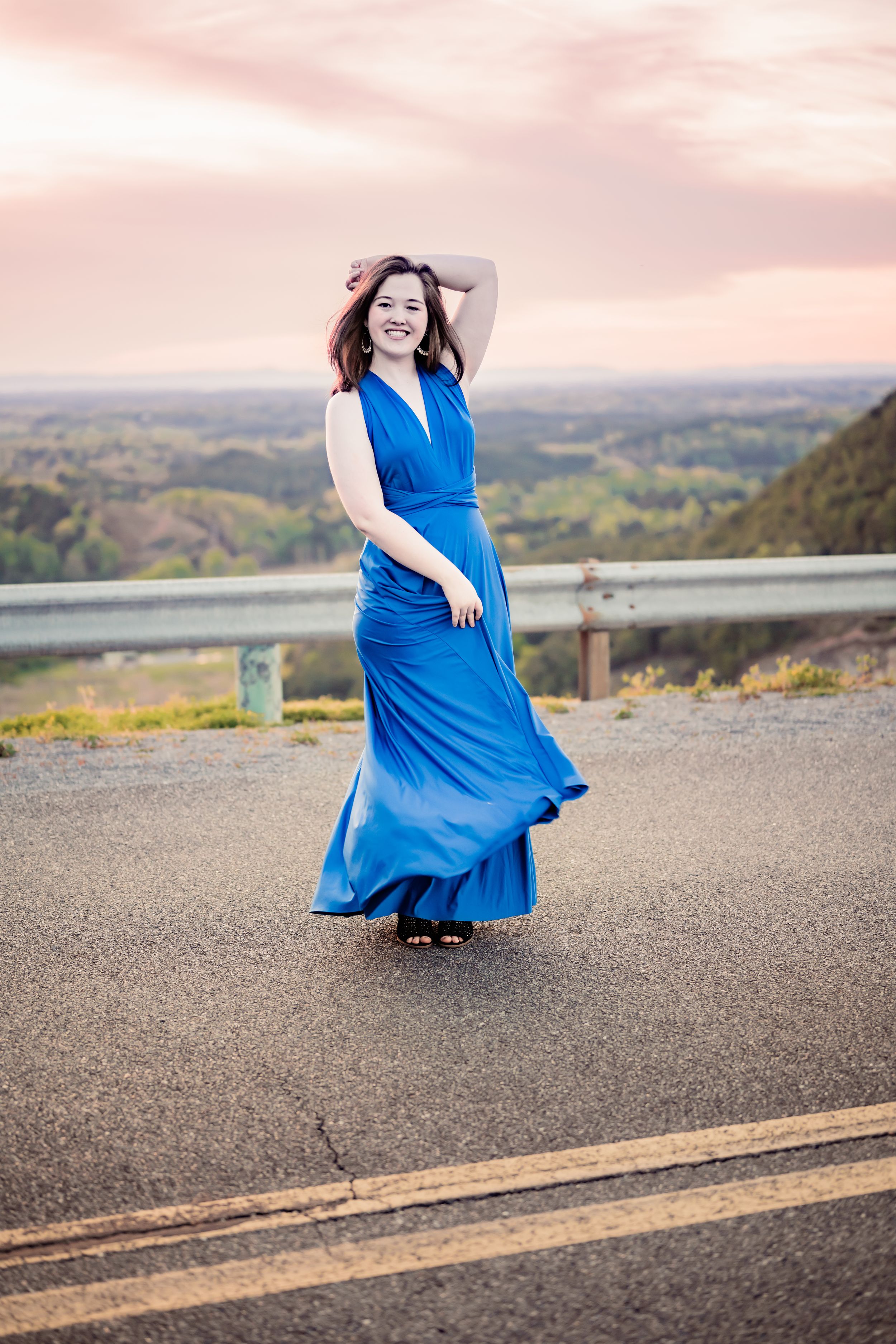 A person in a flowing blue maxi dress poses expressively on a scenic mountain overlook at sunset.