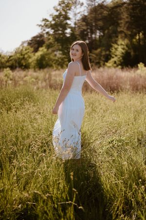 Young woman in white sundress walking through sunlit green meadow with trees in background.