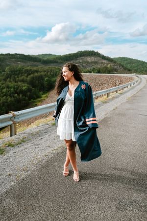 A high school senior in white dress and blue graduation gown poses on mountain road with scenic background.
